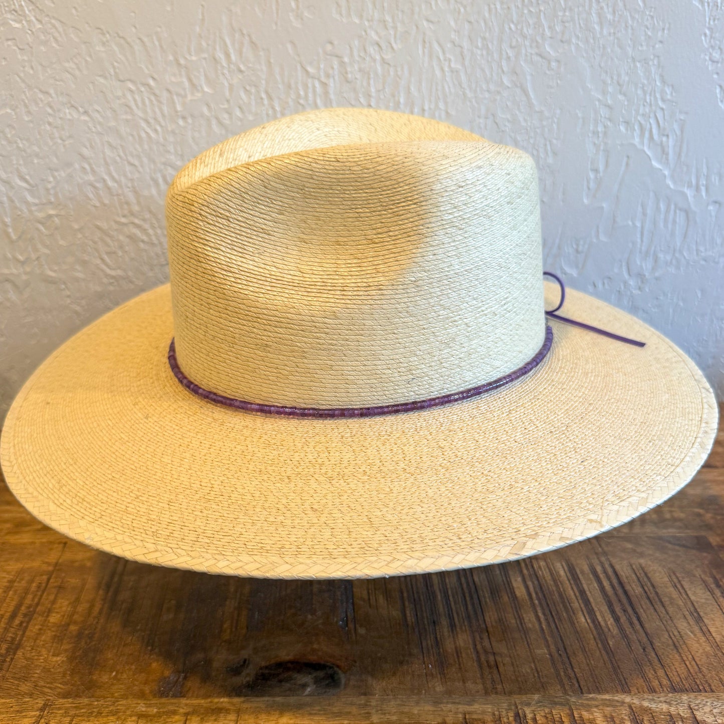 A light beige straw cowboy hat with a dark purple band, displayed on a rustic wooden table against a textured white wall.
