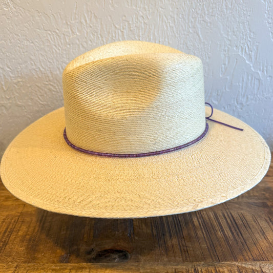 A light beige straw cowboy hat with a dark purple band, displayed on a rustic wooden table against a textured white wall.